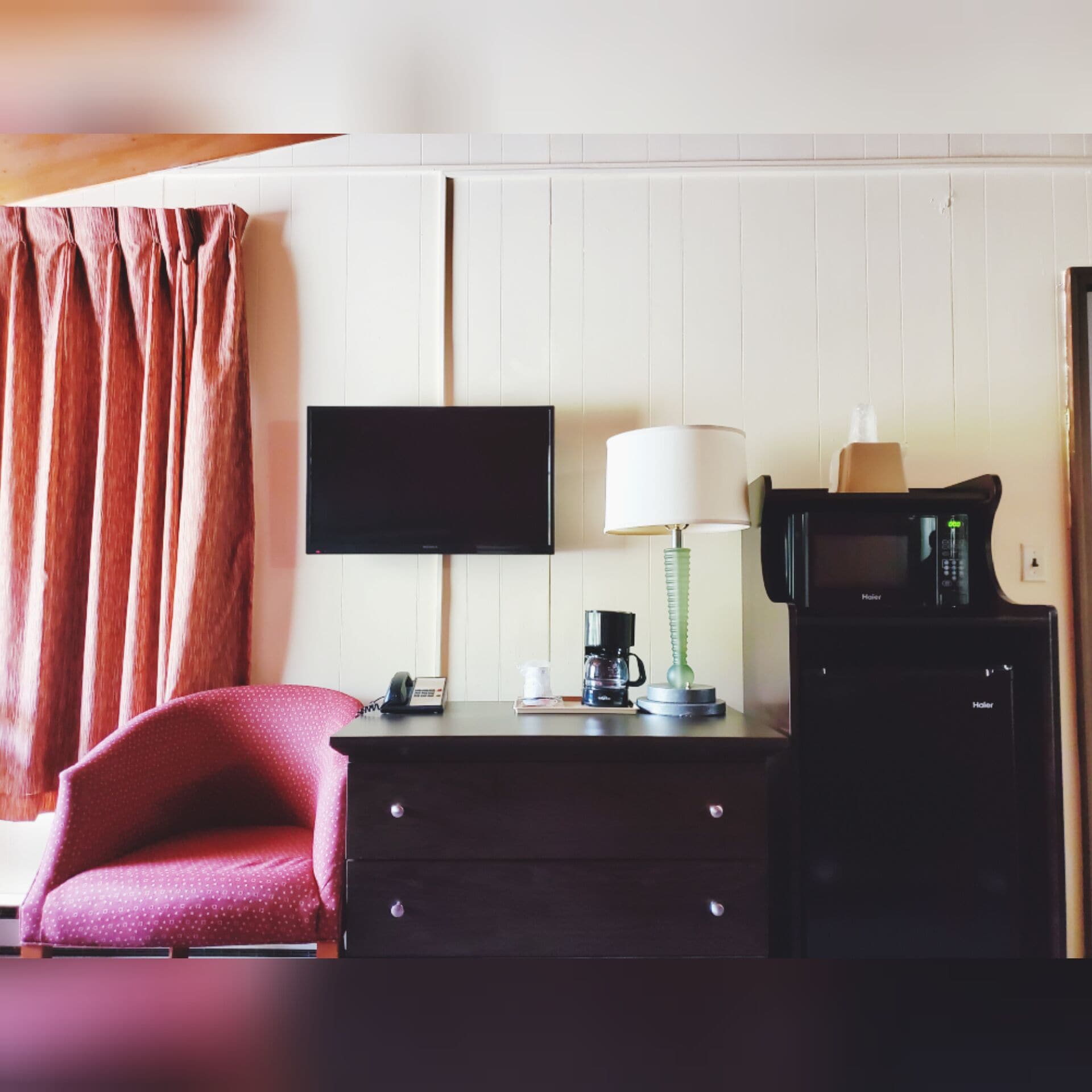A hotel room with a flat-screen TV mounted on the wall, a desk with a lamp, a coffee maker, and a microwave.