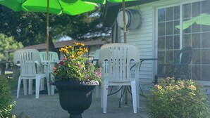 A patio with white plastic chairs, green umbrellas, and a black planter with flowers. - Crossroads Motel (Elkhorn)