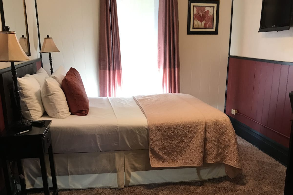 Hotellihuone, jossa on sänky, työpöytä lampun kanssa, televisio ja ikkuna, jossa on verhot.