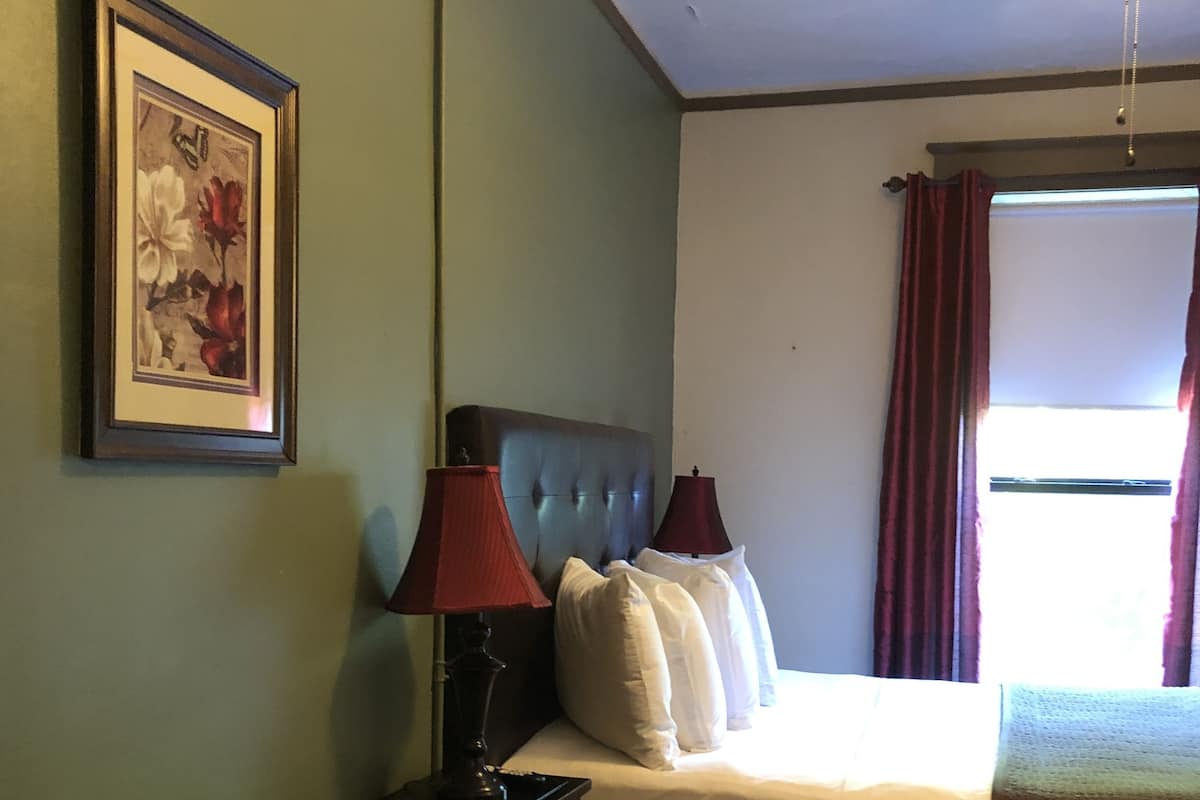 Hotellihuone, jossa on sänky, yöpöytä, lamppu, kattotuuletin, ikkuna verhoilla ja seinällä oleva kehystetty kuva.