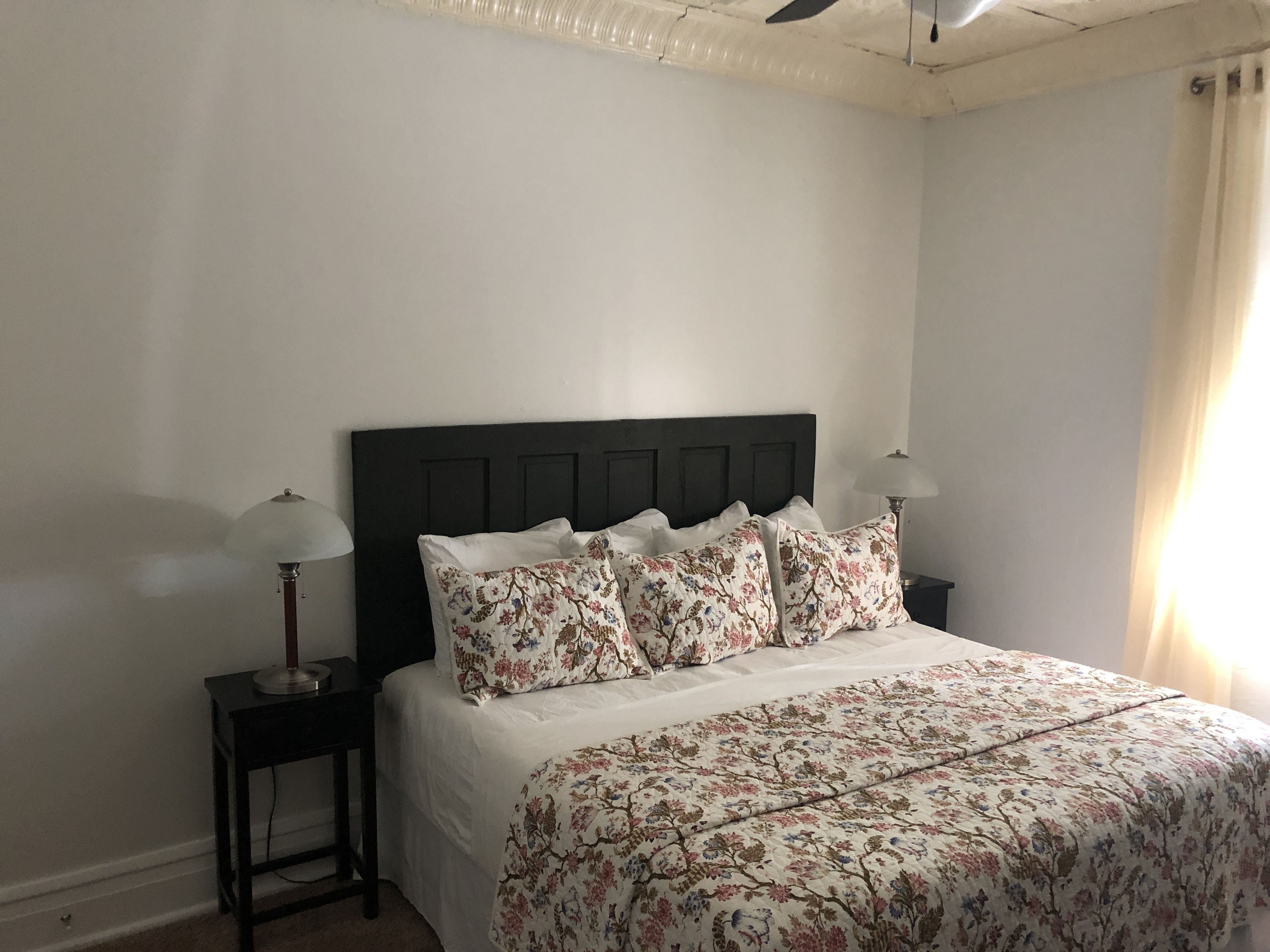 Un dormitorio con cabecera oscura, ropa de cama floral, dos mesitas de noche con lámparas y una ventana con cortinas.