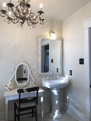 Un baño con tocador blanco, lavamanos pedestal, un espejo grande y una araña.