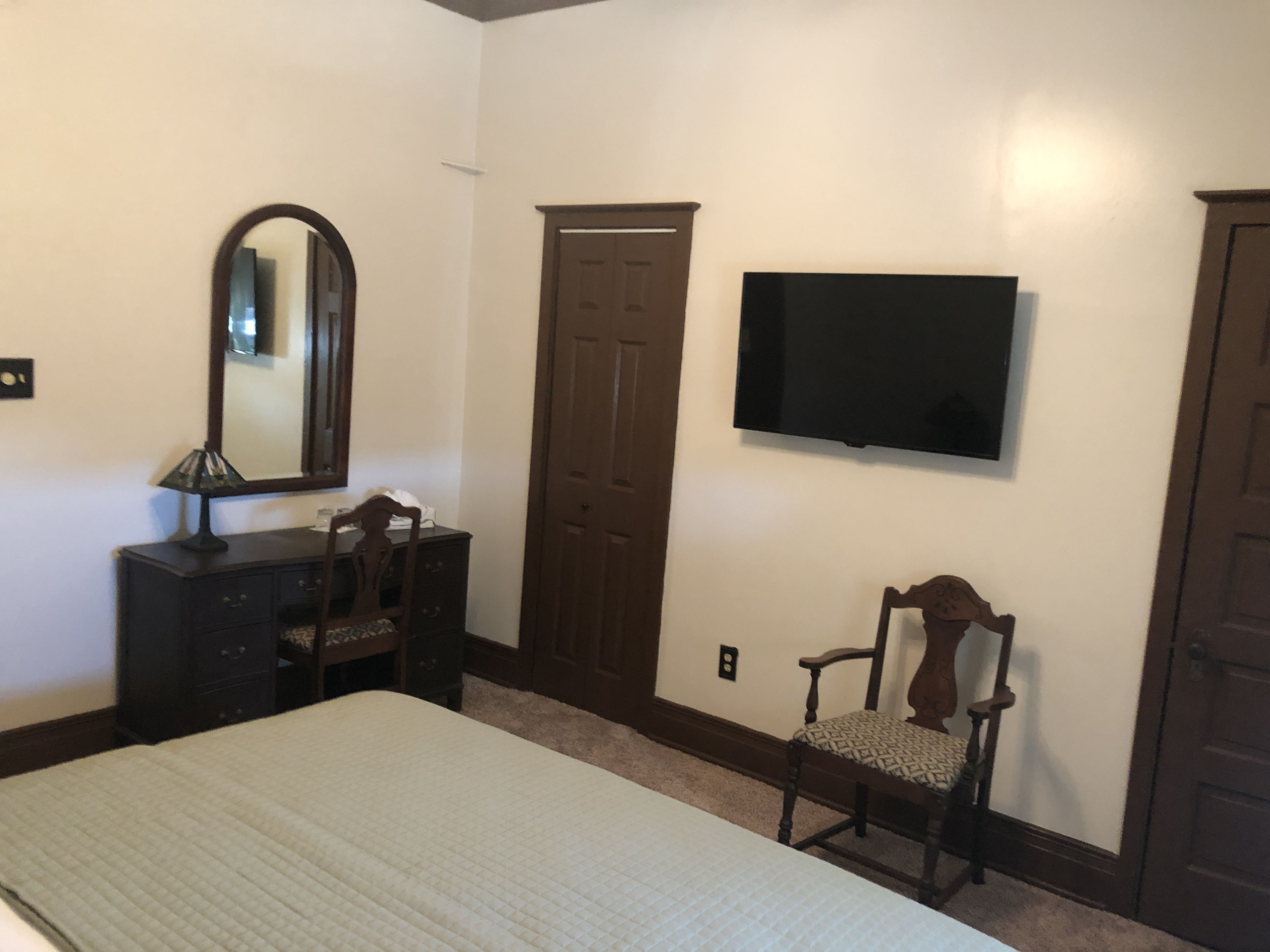 Un dormitorio con cama, cómoda, silla, espejo, armario y televisor montado en la pared.