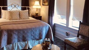 Habitación de hotel con cama, una silla, una mesa, ventilador de techo, ventana con cortinas, lámpara y un cuadro en la pared.