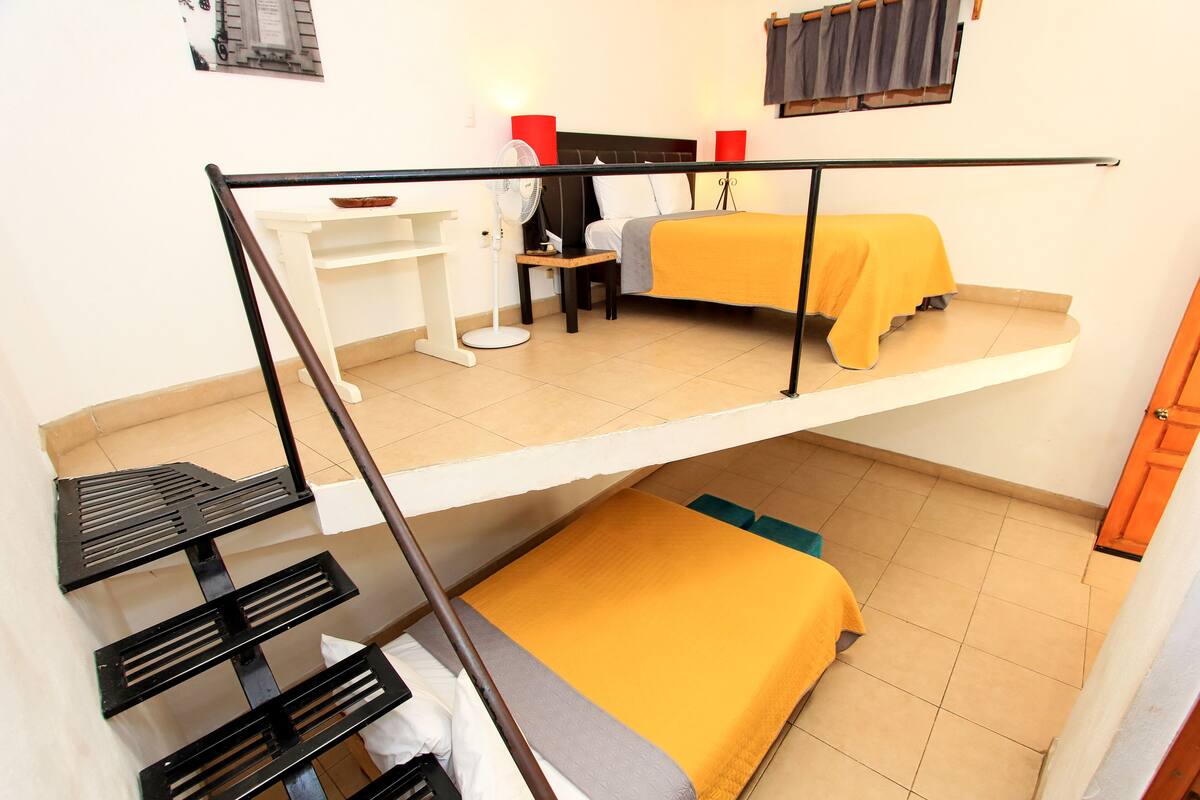 A dormitory room with a bunk bed, a desk with a chair, a small table, and a fan.