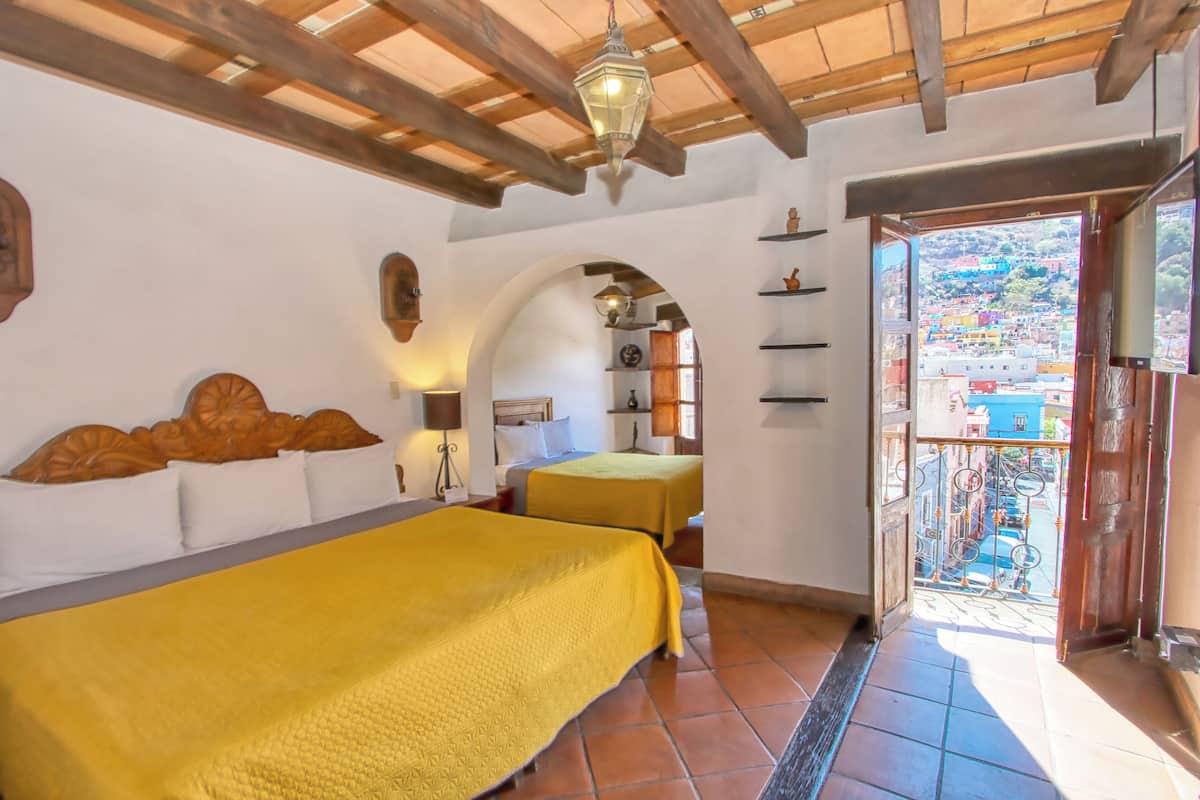 A room with two beds, wooden beams on the ceiling, a balcony with a view of a colorful town, and a door leading outside.