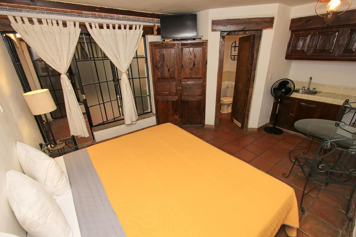 A bedroom with a bed, curtains, a television, a wooden wardrobe, a bathroom, and a kitchenette.