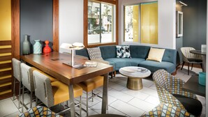 A modern living room with a wooden dining table, blue sofa, patterned chairs, and a large window with a view of the street.