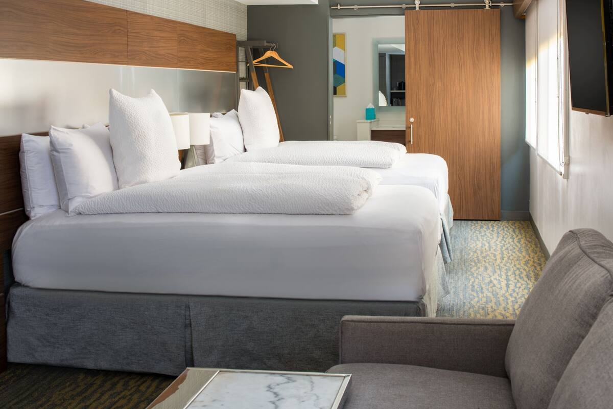 A hotel room with two beds, a sofa, a wooden wardrobe, a mirror, and a framed picture on the floor.
