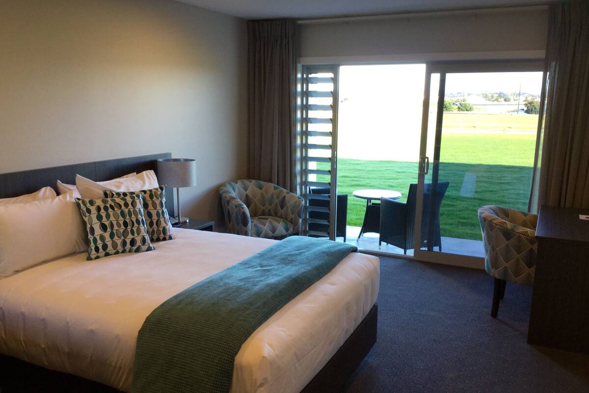A hotel room with a large bed, a desk, two chairs, and a view of a grassy field through a sliding door.