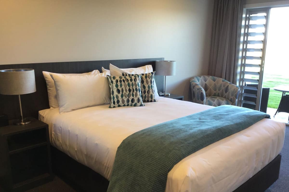A hotel room with a large bed, two bedside lamps, a patterned pillow, a green throw blanket, and a view of the outdoors.