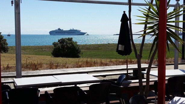 An outdoor seating area with tables and chairs, overlooking a body of water with a cruise ship in the distance. - East Pier Hotel (Napier)