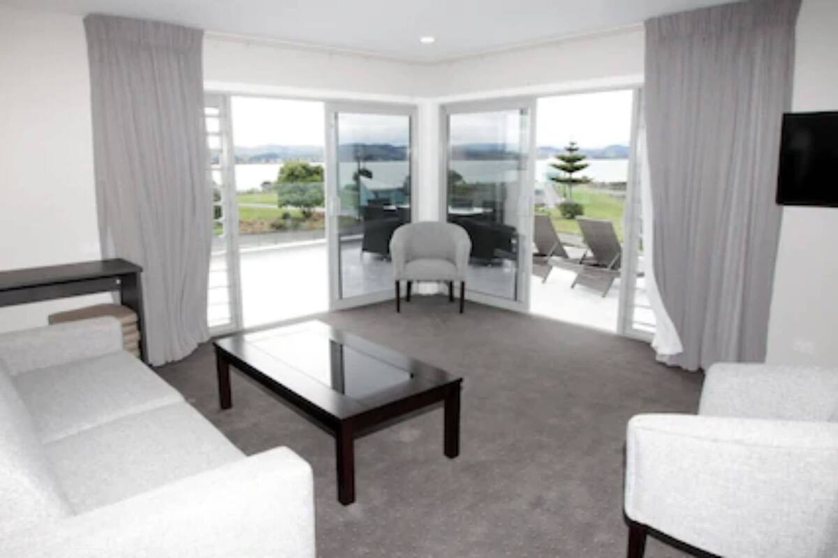 A modern hotel room with a sofa, armchair, and a table. There is a balcony with a view of the landscape.
