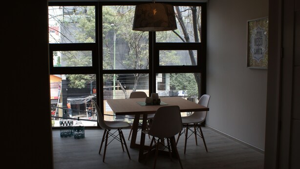 Un comedor con una mesa y sillas, ventanas amplias con vista a una obra en construcción, y una lámpara colgante.