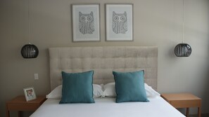 Une chambre à coucher avec une tête de lit capitonnée, deux coussins turquoise, un couvre-lit blanc et deux œuvres d’art encadrées accrochées au mur au-dessus du lit.