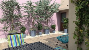Un patio al aire libre con una mesa negra y dos sillas, rodeado de plantas en macetas y un muro rosa.