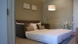 Una cama bien hecha con un edredón blanco, un cabecero marrón y almohadones decorativos. Se observa una lámpara de pie con pantalla blanca y un jarrón con flores blancas al lado.