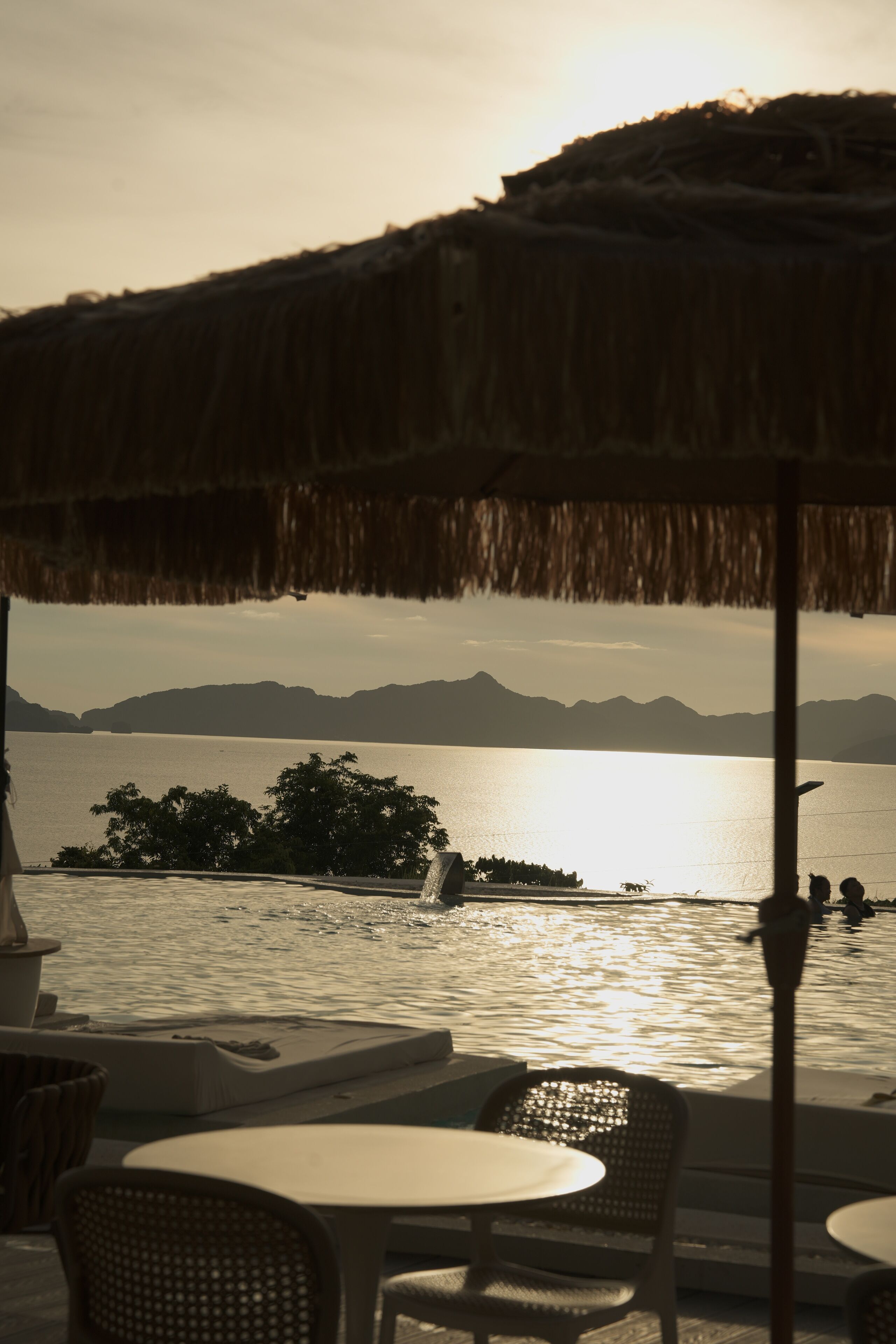A seaside patio with thatched umbrellas, tables, and chairs overlooking a calm body of water and distant mountains.