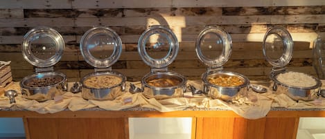 A buffet with various food items in metal containers, set against a wooden wall with slatted ceiling lights.