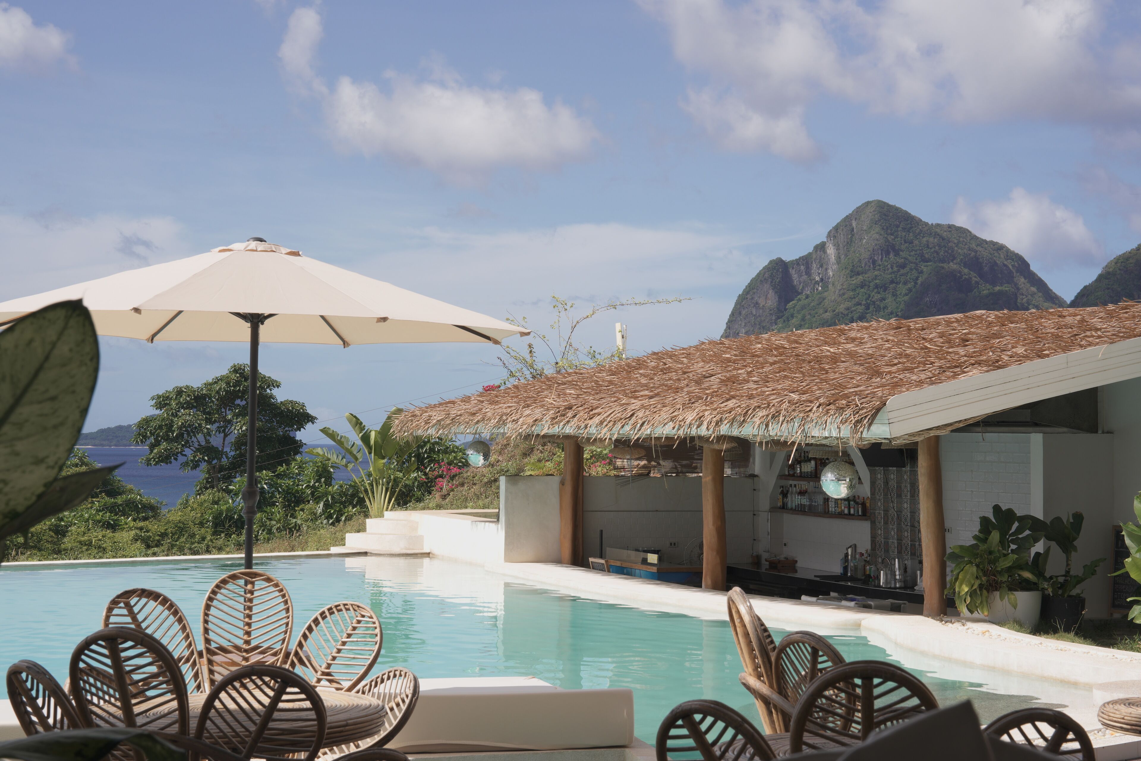 A poolside area with lounge chairs and an umbrella, a bar, and a view of the ocean and mountains.