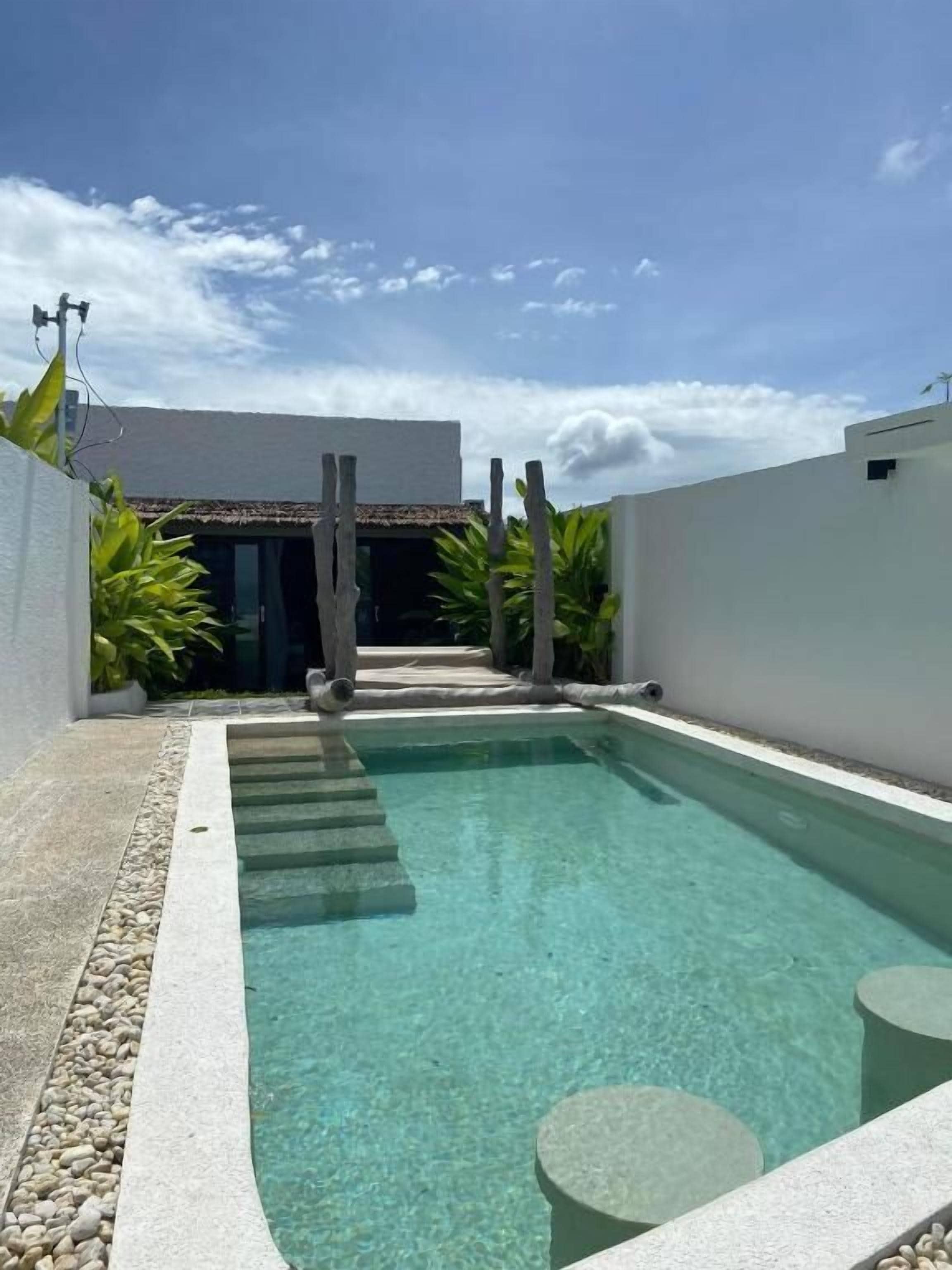 Una piscina con agua cristalina, rodeada de muros blancos y un pequeño patio con escalones que conducen a un edificio.