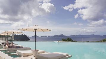 Zona junto a la piscina con sillones de descanso y sombrillas, con vista a un mar azul cristalino y montañas a lo lejos.