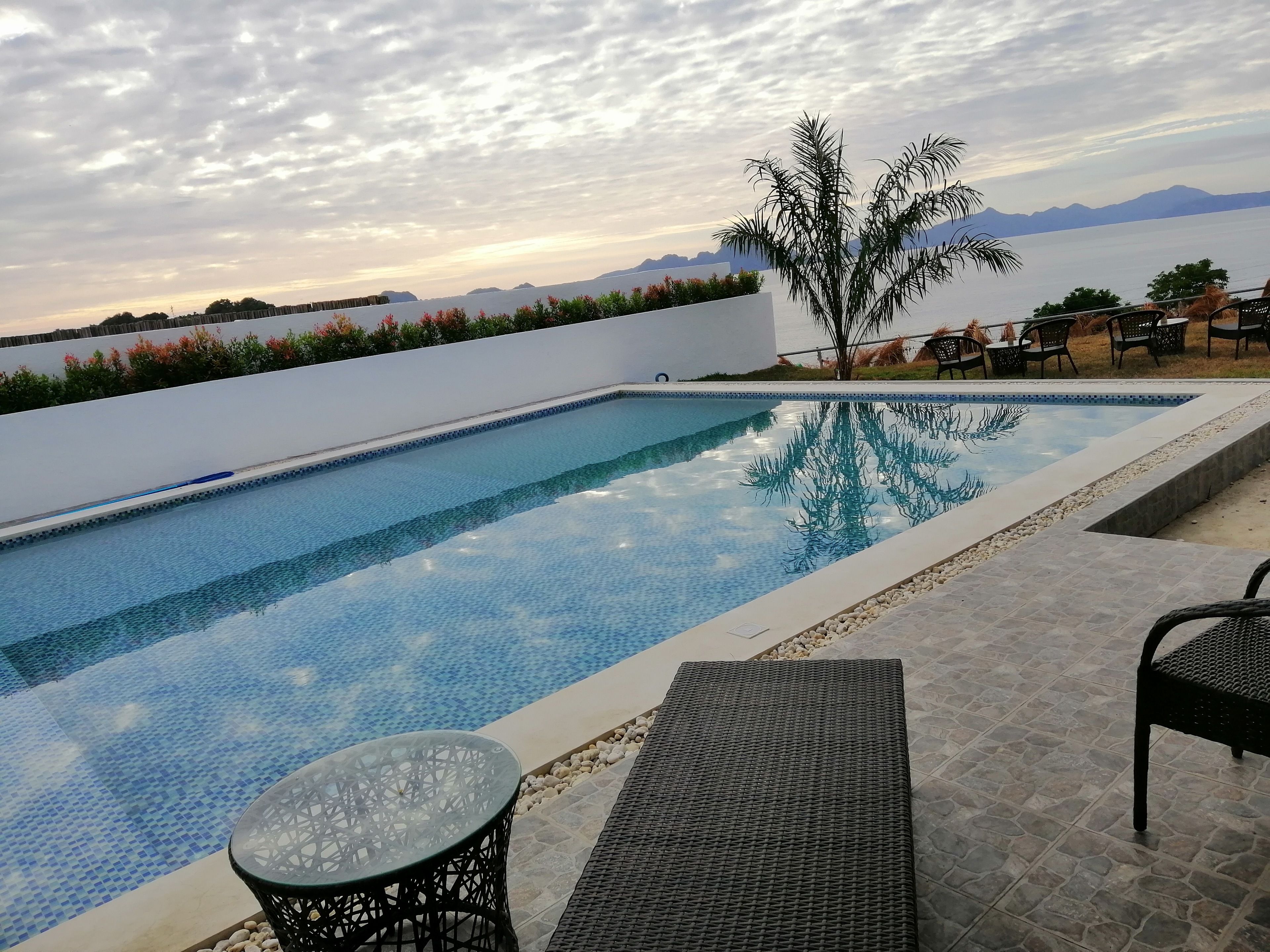 Une piscine rectangulaire à l’eau d’un bleu limpide, entourée d’un mur blanc et d’une terrasse carrelée. On y trouve deux chaises en osier noir et une table en verre, près de la piscine.
