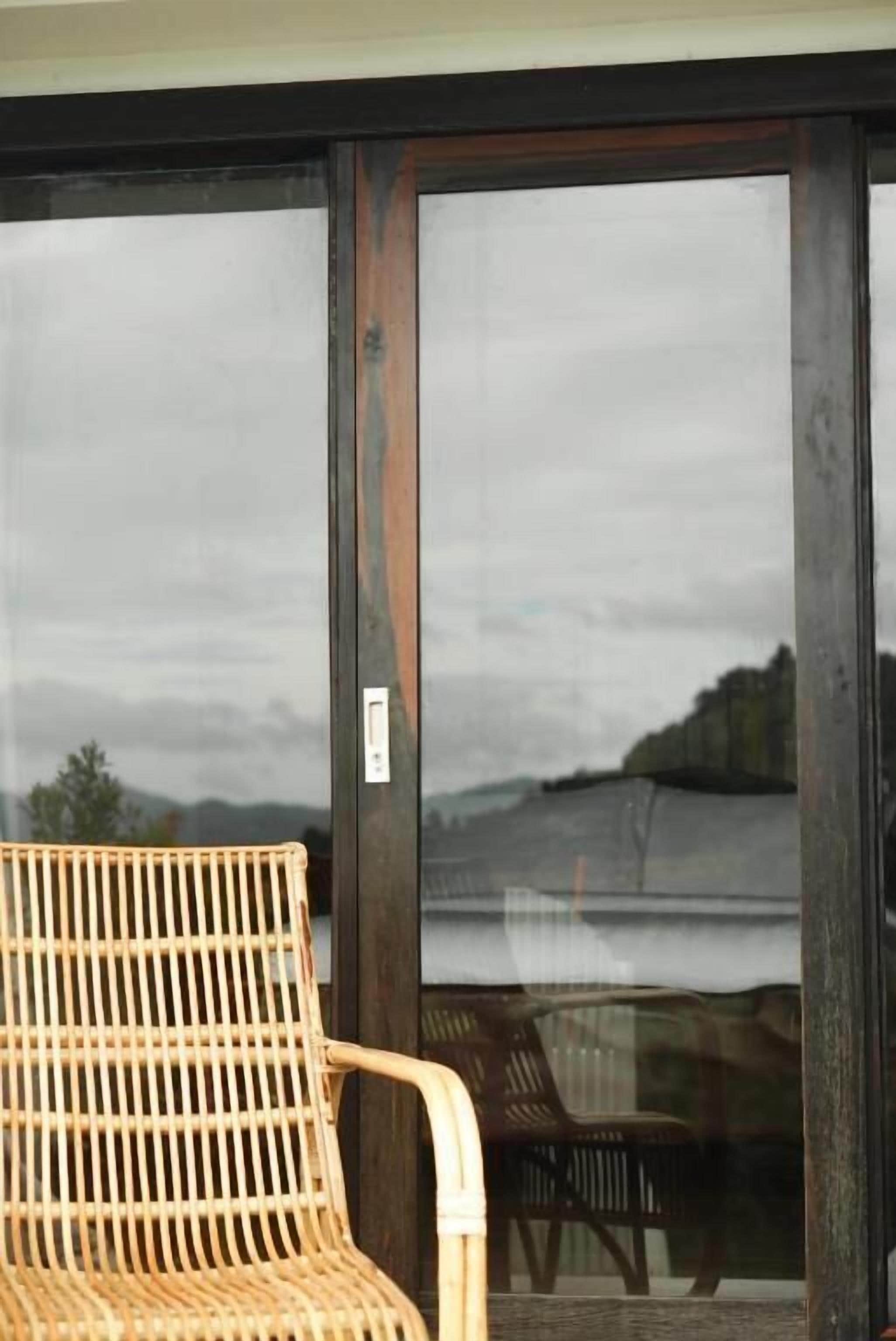 Une chaise en osier est placée devant une porte-fenêtre coulissante, offrant une vue sur un lac et des collines.
