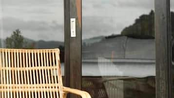 Une chaise en osier est placée devant une porte-fenêtre coulissante, offrant une vue sur un lac et des collines.