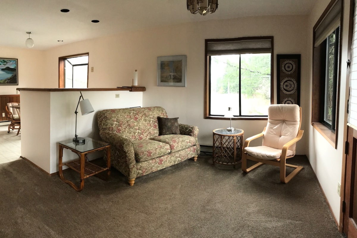 Ein Wohnzimmer mit floralem Sofa, einem Holzstuhl, einem kleinen Tisch und einer Lampe. Es gibt ein Fenster mit Blick auf Bäume und eine Wand mit dekorativen Tellern.