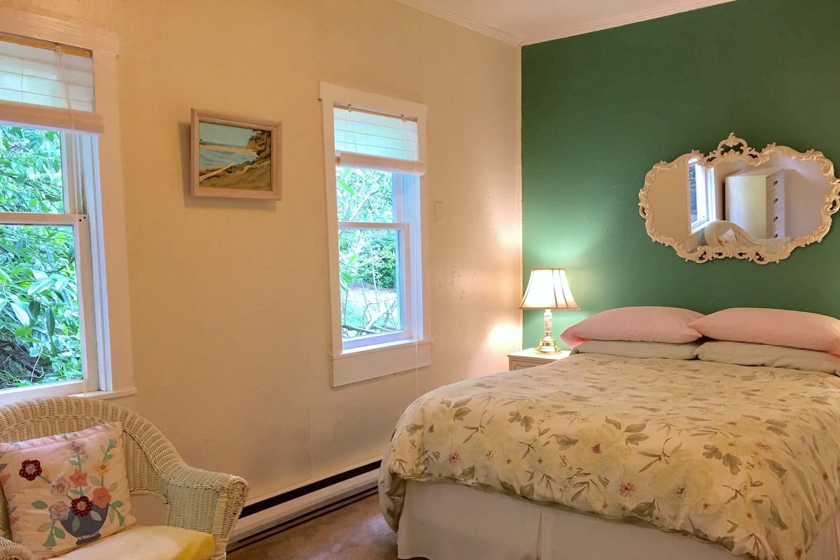 Ein Schlafzimmer mit einem Bett, einem Sessel, einem Nachttisch, einer Lampe, einem Spiegel und zwei Fenstern.