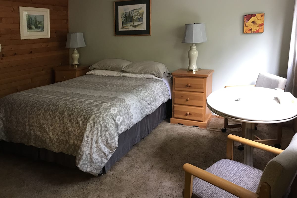 Ein Schlafzimmer mit einem Bett, zwei Nachttischen mit Lampen, einem Sessel, einem kleinen runden Tisch und einem Bild an der Wand.