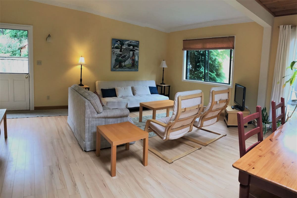 Ein Wohnzimmer mit einer Couch, einem Holztisch, einem Sessel, einem Fernseher und einem Fenster mit Blick ins Grüne.