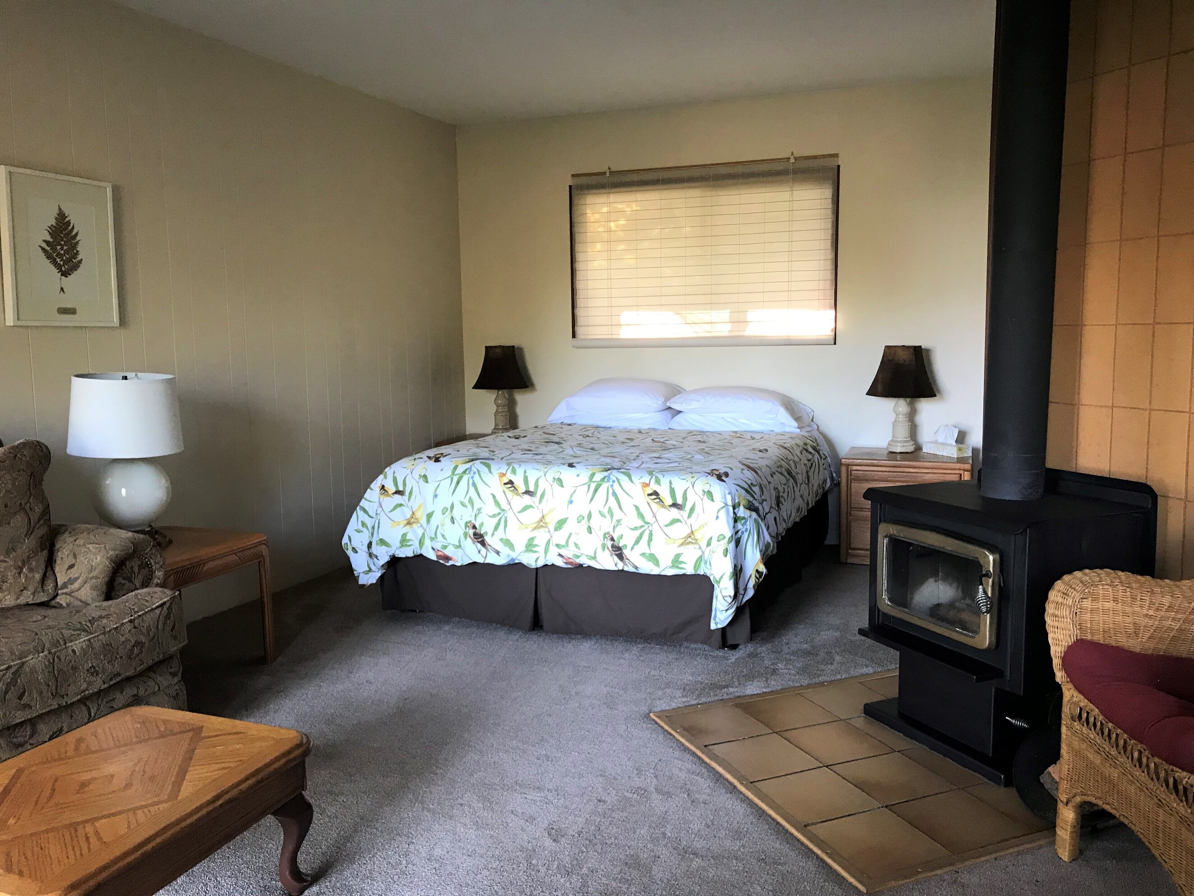 A cozy bedroom with a bed, two bedside lamps, a wooden nightstand, a wicker chair, a small wooden table, and a wood-burning stove.