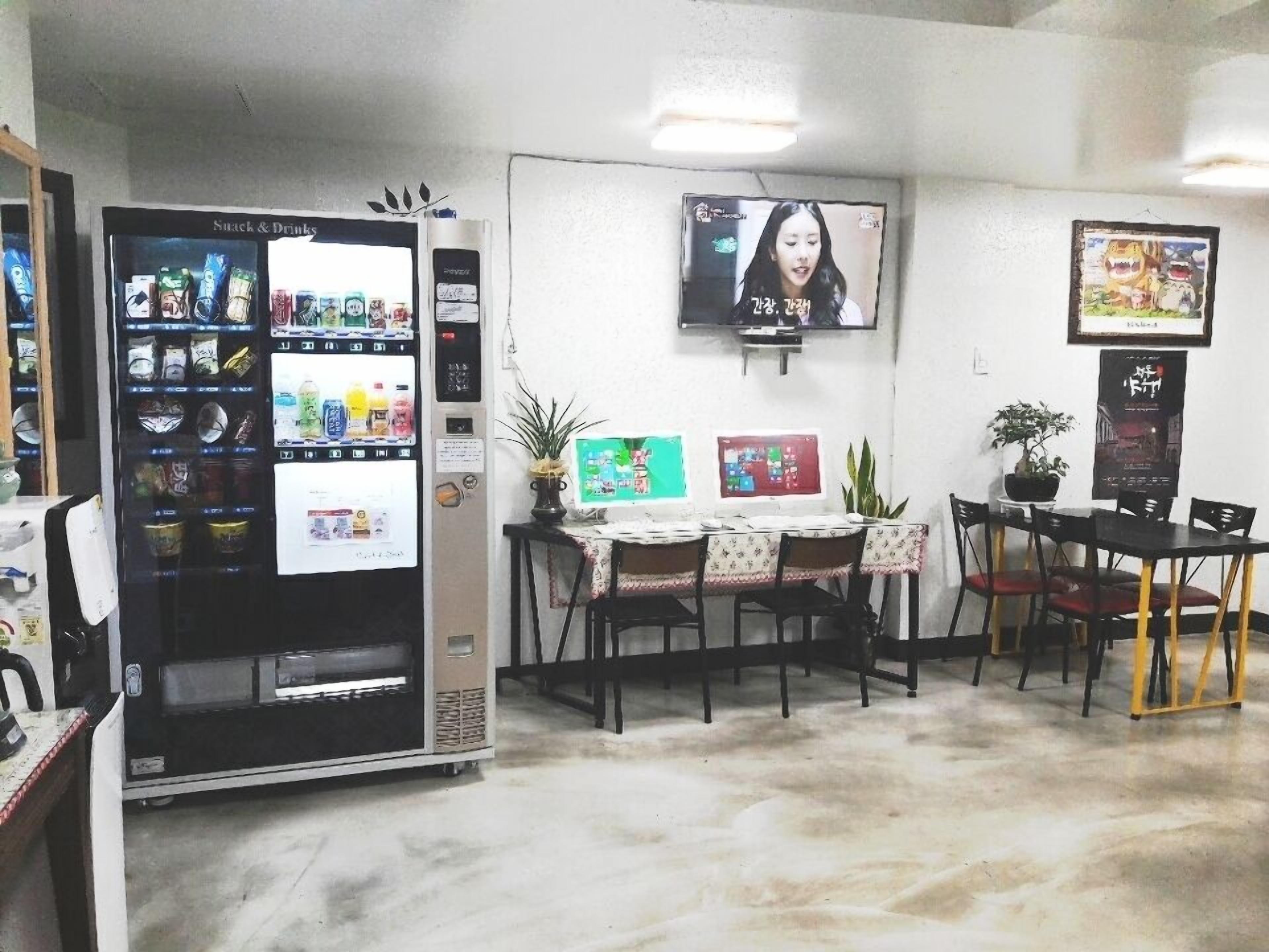 A room with a vending machine, a TV mounted on the wall, a table with chairs, and a potted plant.