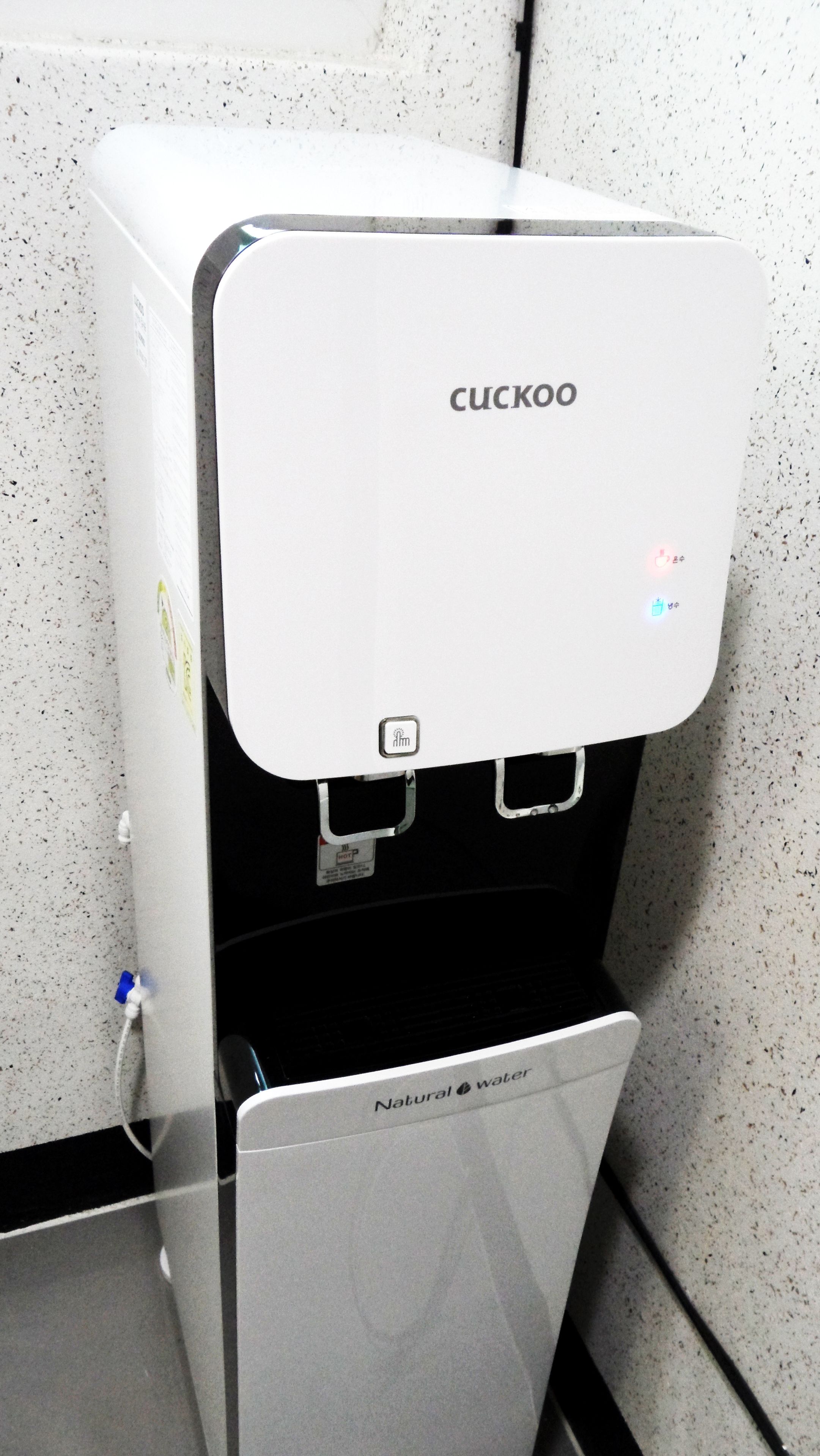 Un dispensador de agua Cuckoo con etiqueta 'Natural Water', una toma de corriente y un cartel de 'No Fumar'.