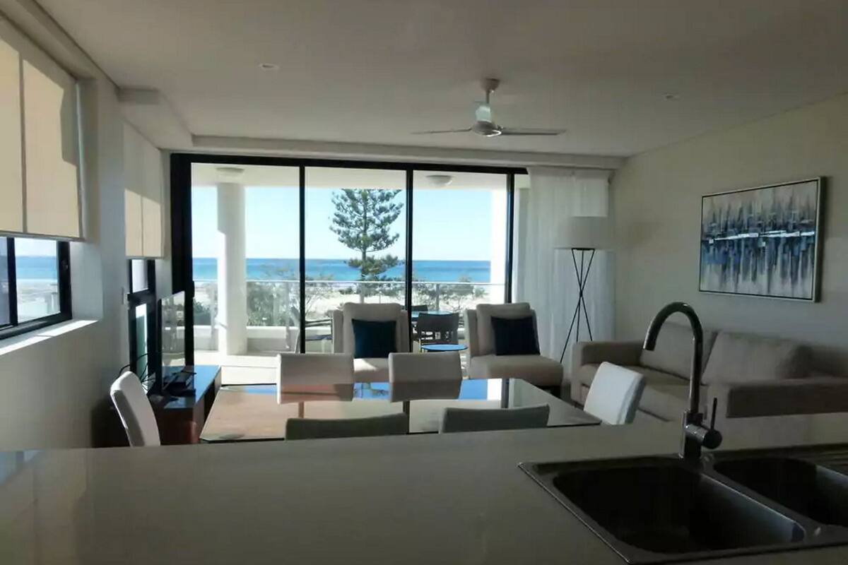 Una cocina moderna con una isla grande, un comedor con vista al océano y una sala de estar con ventilador de techo y obras de arte.