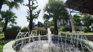 Una fuente circular con múltiples chorros, rodeada de palmeras y una estructura cubierta.
