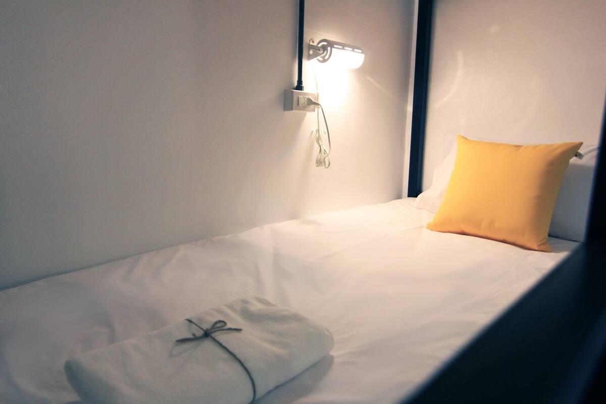 Un lit simple avec un matelas blanc, un oreiller jaune et une couverture blanche attachée avec une ficelle.