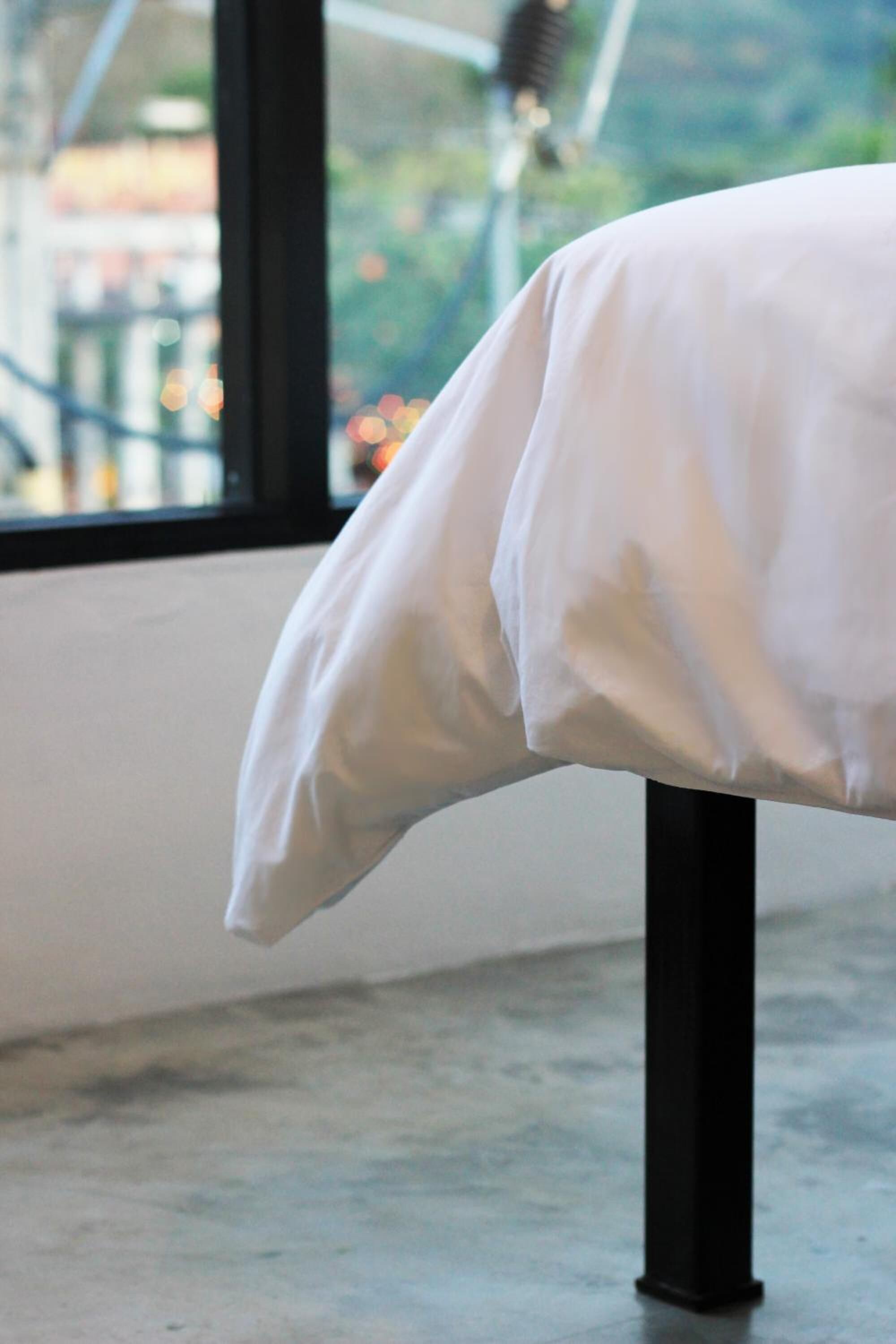 A bed with a white sheet, a black metal frame, and a view of a cityscape through a window.