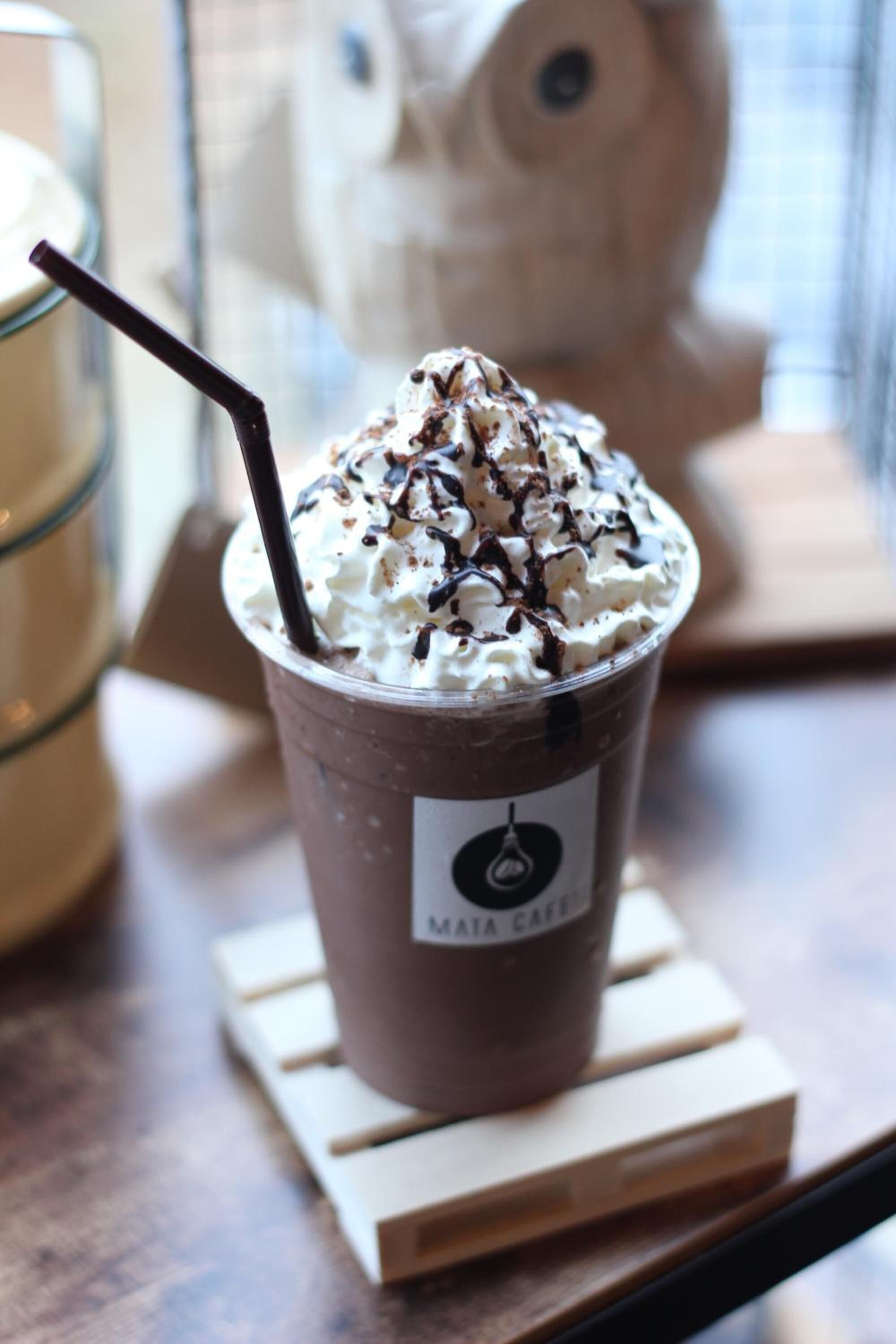 A cup of chocolate milkshake with whipped cream and chocolate shavings, labeled "Maya Cafe," on a wooden coaster.