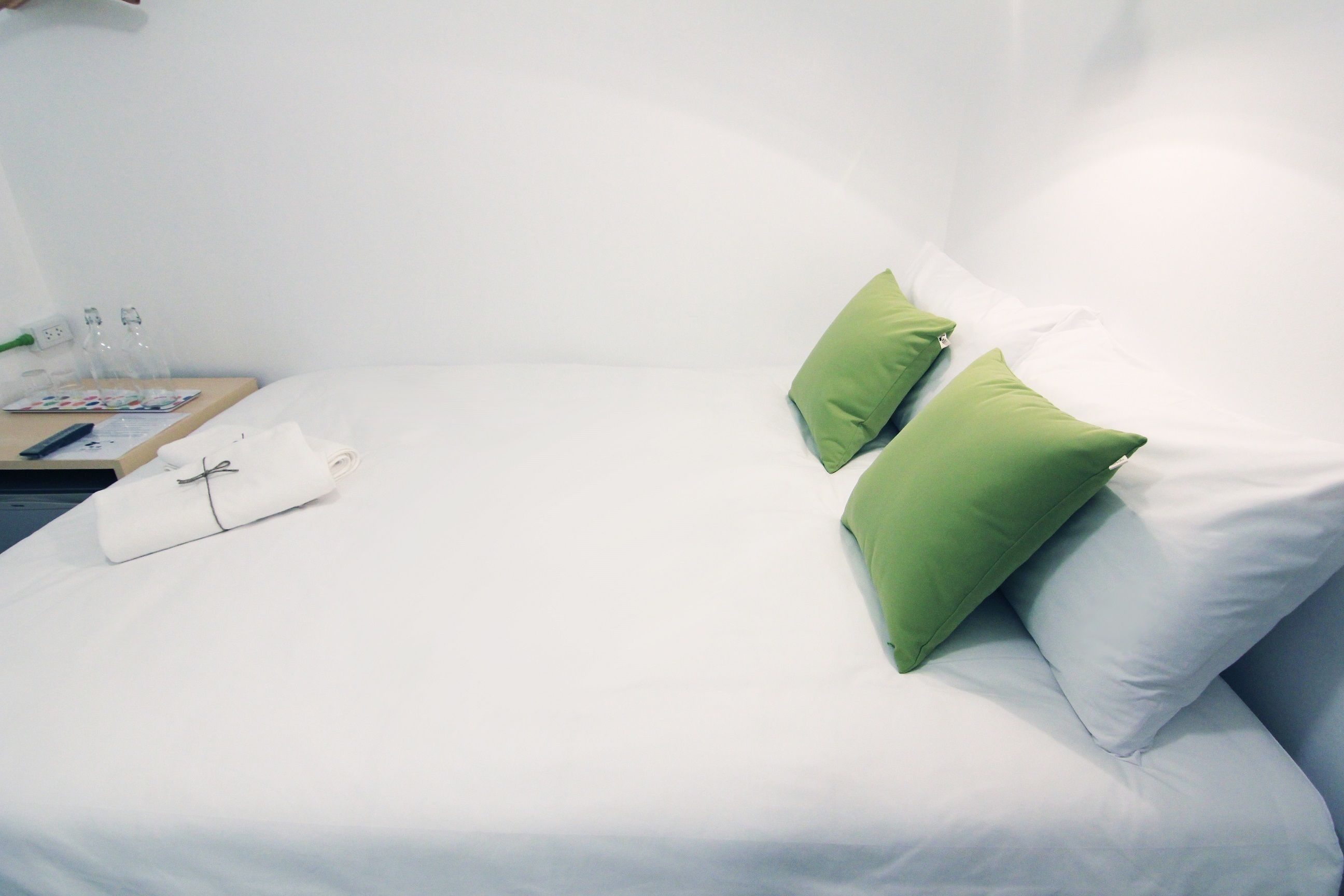 Un lit bien fait, avec du linge de lit blanc et deux oreillers verts, une petite table avec une télécommande, une serviette pliée et quelques bouteilles à côté.