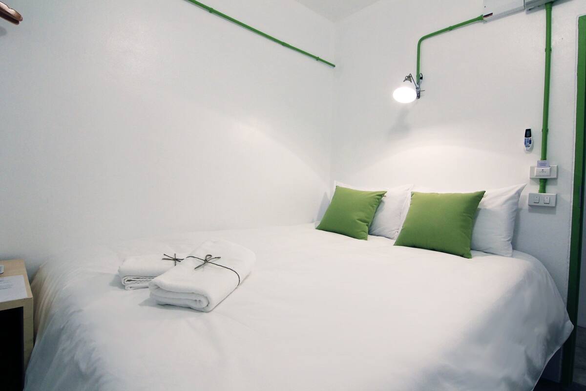 Un lit bien fait, avec du linge de lit blanc et des coussins verts, une table de chevet avec une lampe et un panneau de commande fixé au mur.