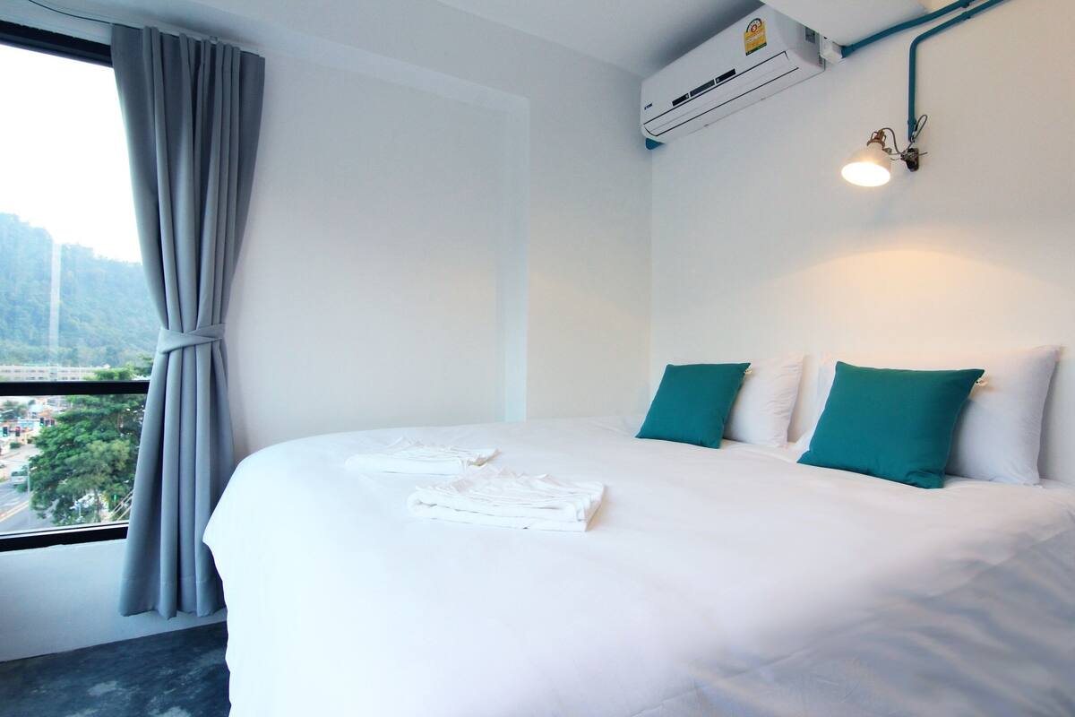 Une chambre d’hôtel avec un lit bien fait, une fenêtre avec des rideaux, un appareil de climatisation et un luminaire fixé au mur.