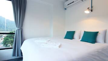 A hotel room with a neatly made bed, a window with curtains, an air conditioning unit, and a wall-mounted light fixture.