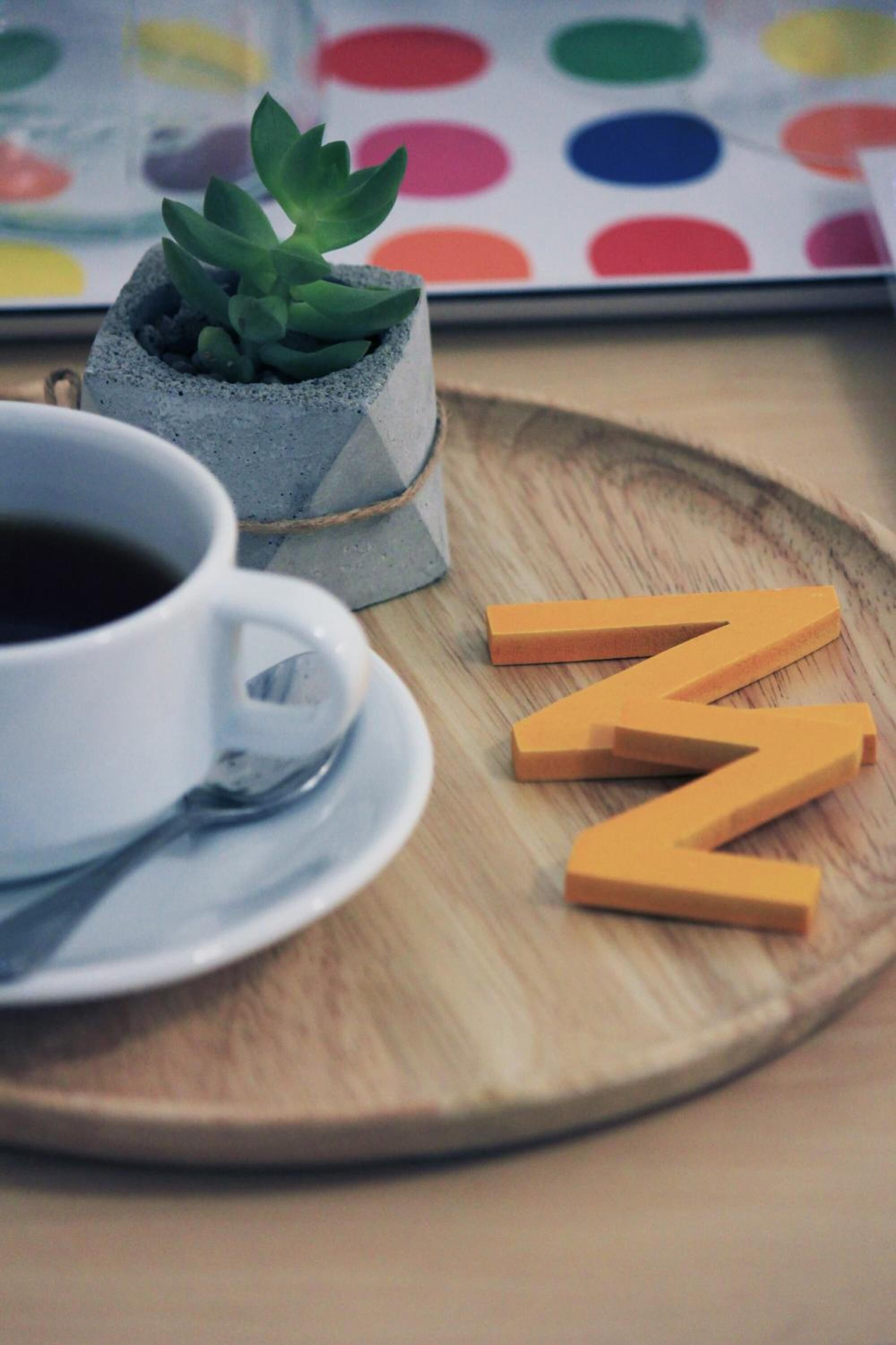 Una tassa de cafè blanca sobre un plat, una petita planta en test i blocs de lletres de fusta sobre un safata de fusta.