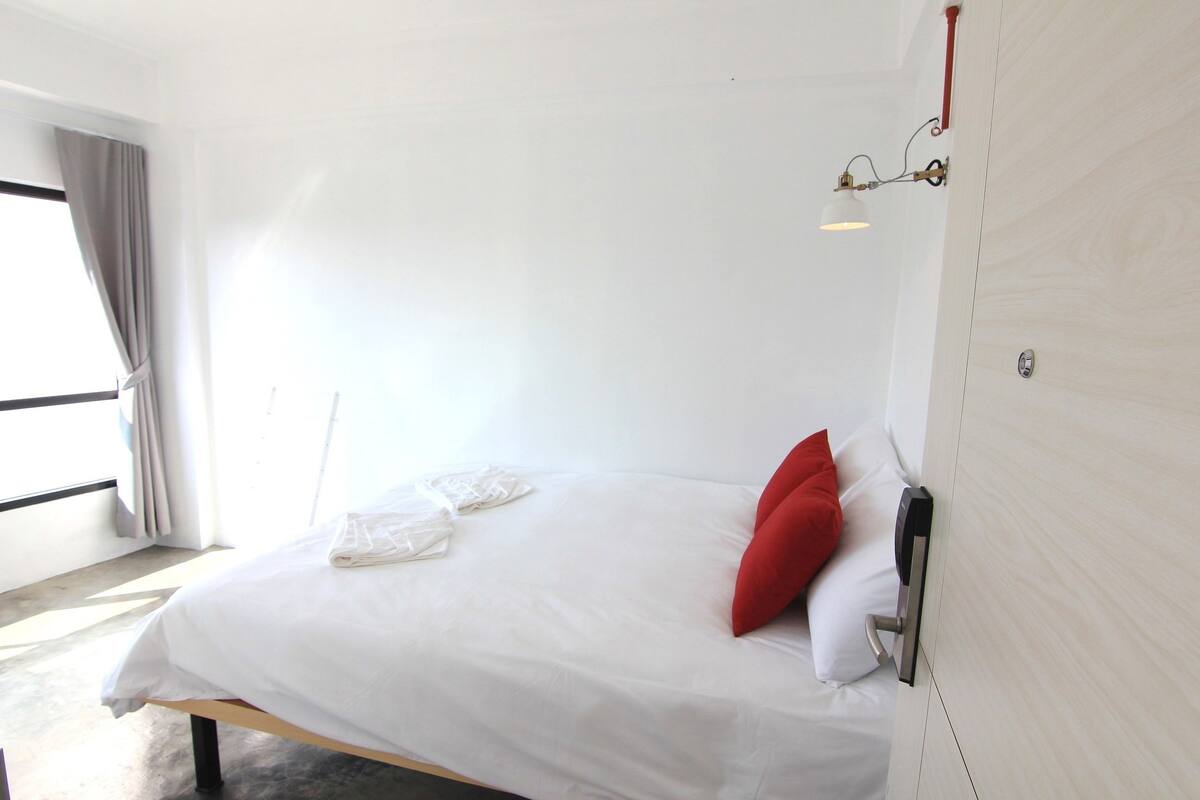 Un lit bien fait, avec une literie blanche et un oreiller rouge, une table de chevet avec une lampe, et une fenêtre avec des rideaux.