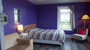 Een slaapkamer met een bed, nachtkastje, lamp, stoel en een raam met gordijnen.