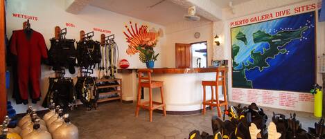Tienda de buceo con trajes de neopreno, chalecos BCD y equipo de escafandra. En la pared, se aprecia un mapa de los puntos de buceo de Puerto Galera.