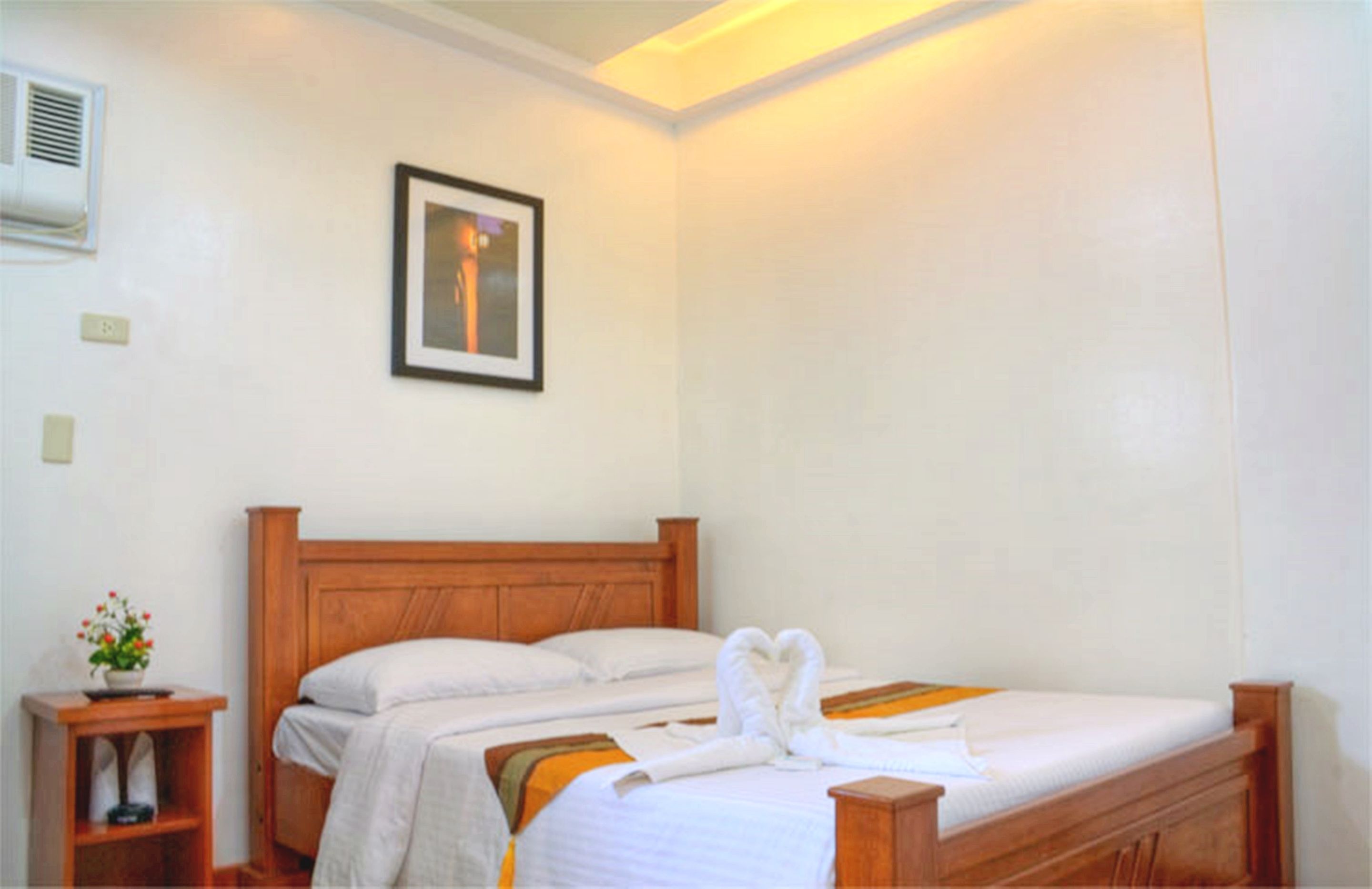 Una habitación de hotel con dos camas individuales, un cuadro enmarcado en la pared, una mesita pequeña con un florero y una mesita de noche con una lámpara.